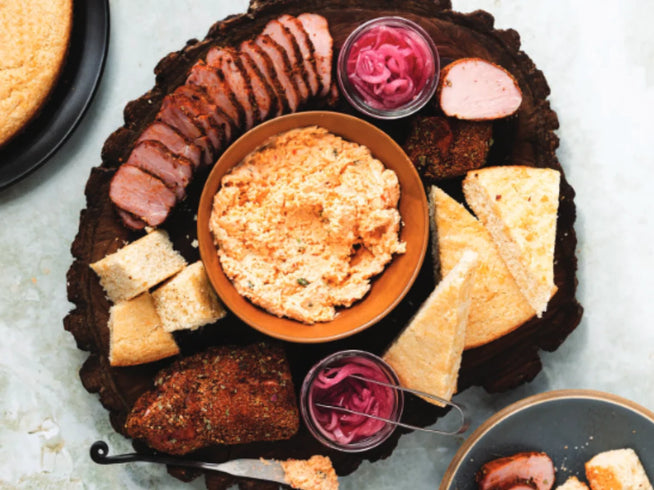 Overhead view of a wooden serving board with a bowl of pimento cheese, sliced pork tenderloin, pickled red onions, and wedges of cornbread.
