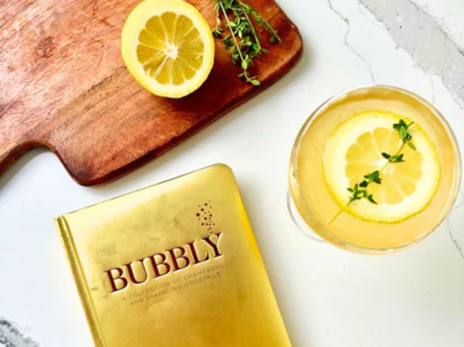 Gold 'Bubbly' book next to a glass of lemonade with lemon slices and thyme on a white surface.