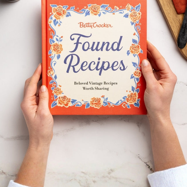 Book titled 'Found Recipes' by Betty Crocker held by hands on a light surface.